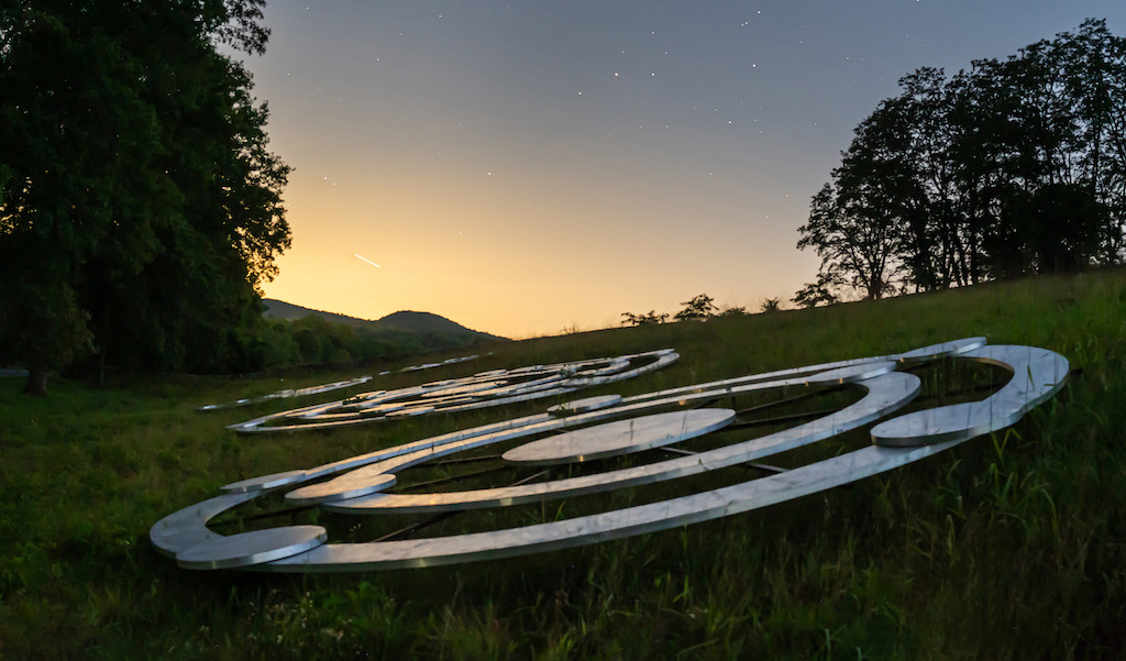 In the foreground, a sculptural installation of large concentric metallic rings lies on a grassy hillside. To the left, tall trees cast dark silhouettes, while to the right, the edge of a wooded area is visible against the horizon. In the background, rolling hills fade into the distance under a gradient sky that transitions from warm yellow near the horizon to deeper blue tones higher up.