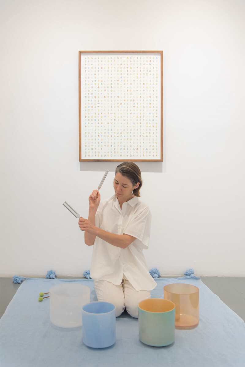 Dressed in a white shirt and pants, Kiko is seated on a blue blanket surrounded by four sound bath bowls of different sizes and colours with two tuning forks in her hands. On the white wall behind Kiko is a framed artwork consisting of hundreds of miniature multicolour shapes against a white surface.