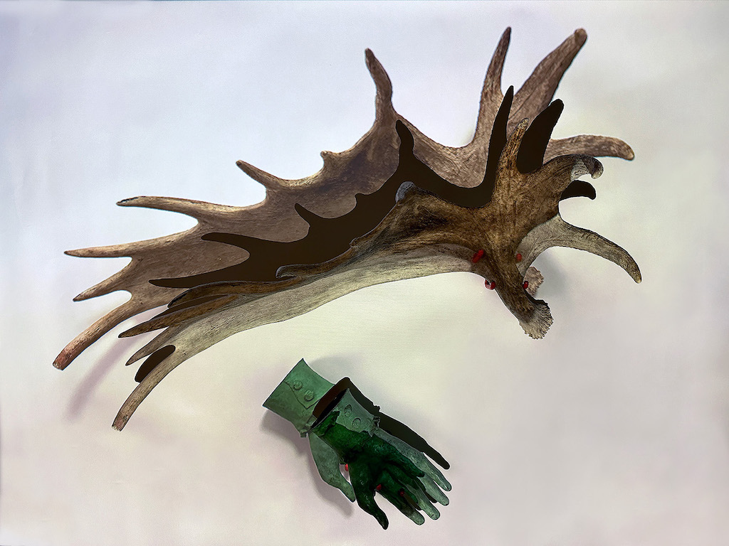 This image showcases two distinct objects arranged against a clean white background. At the top, a large moose antler with a rugged texture and branching points extends outward. Below the antler, a green hand lies flat, palm facing upward, adorned with a two-buttoned cuff.