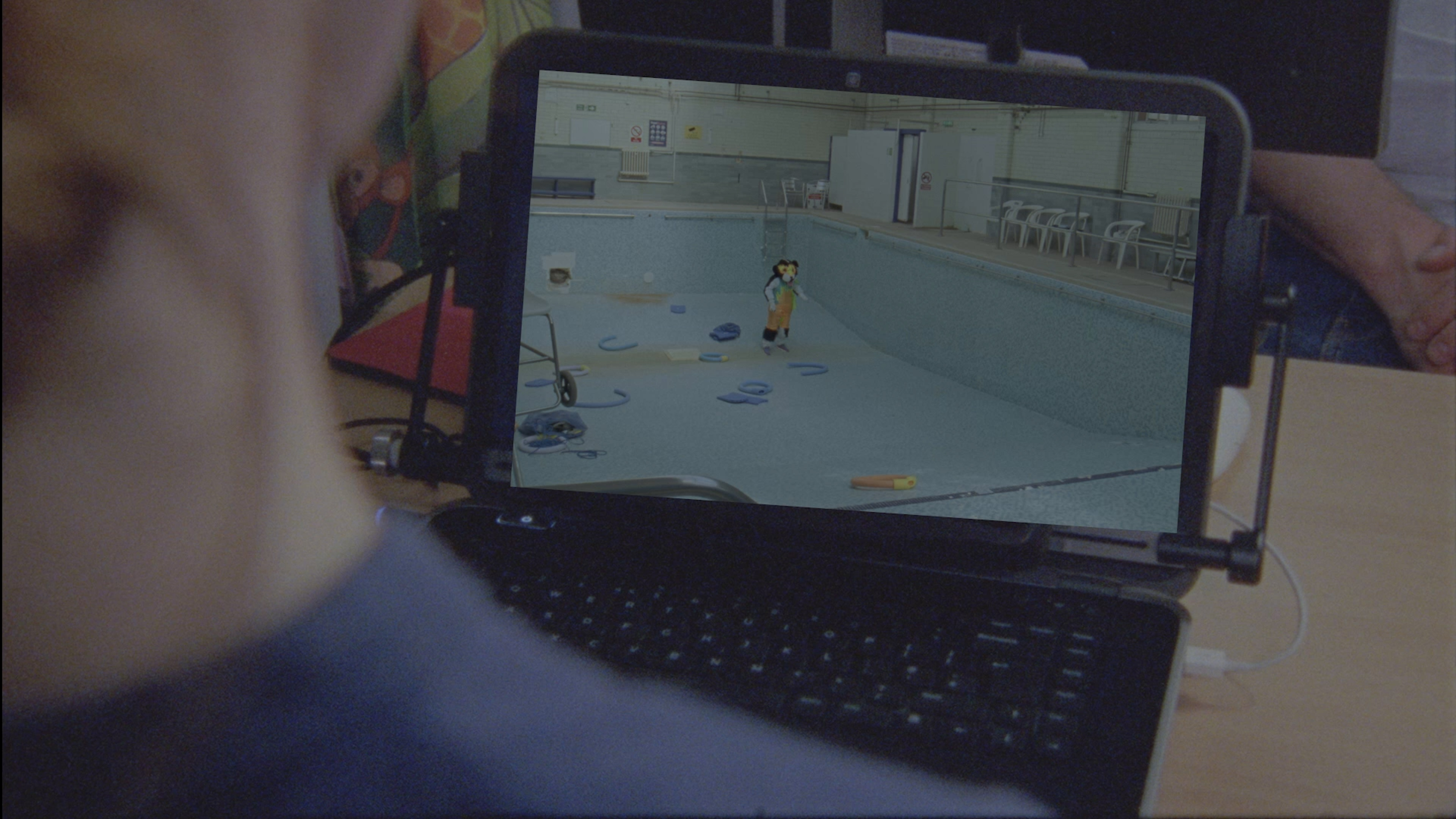 A close-up of a laptop screen with a video playing, showing an empty, drained indoor swimming pool, surrounded by plastic chairs lined up against tiled walls. In the pool, there is a person wearing a large cartoonish animal costume standing alone amidst scattered swimming equipment like foam floats and pool noodles. In the foreground, part of a person's head and shoulder is visible, slightly out of focus, as they watch the screen. On the desk surrounding the laptop, there are various items, including cables and folders.