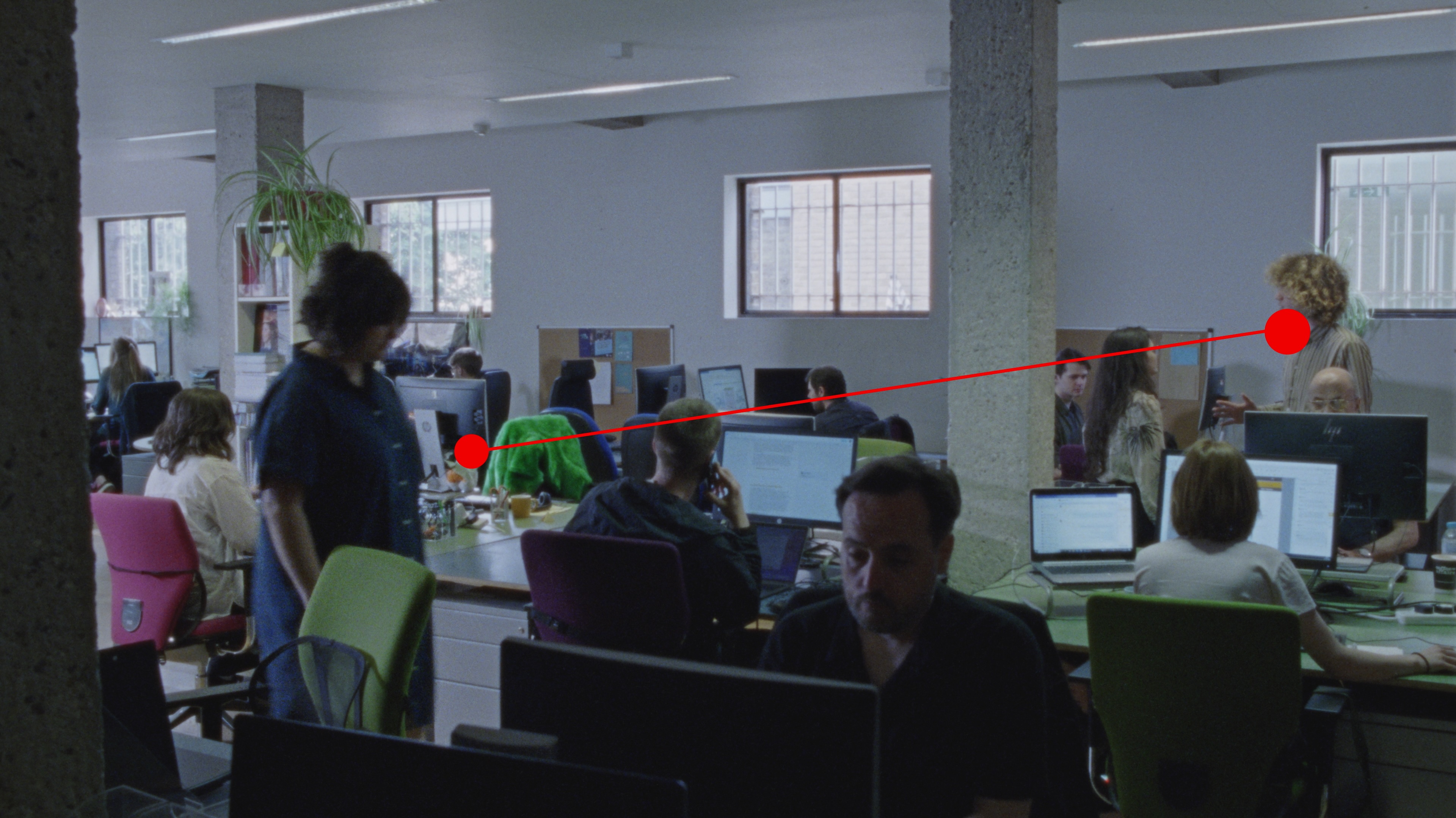 An open-plan office space with multiple people engaged in various activities, such as working at computers, talking on the phone and in conversation with each other. A red line with dots at both ends cuts horizontally across the image, representing the gaze of an eye-tracking test participant.