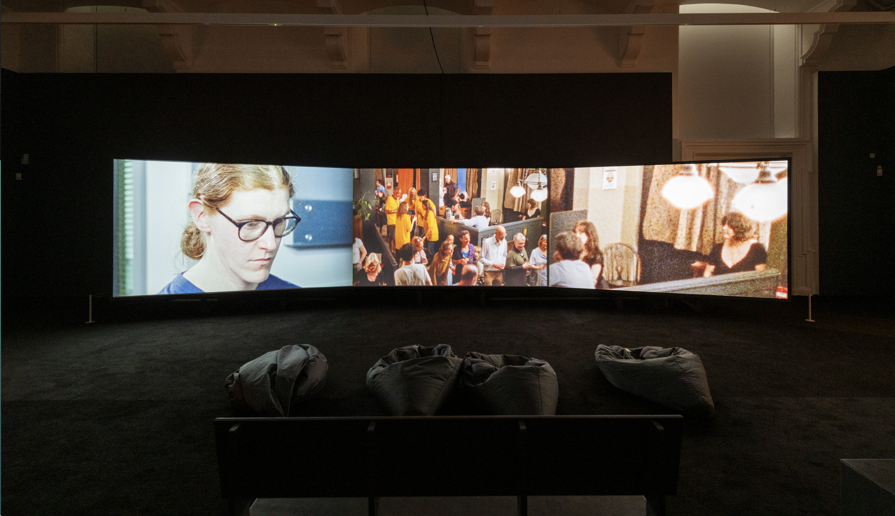 A three-screen video installation in a large, carpeted room with four beanbags and backed benches. From left to right, the screens show: (1) A close-up of person, with tied-back hair and wearing black-rimmed glasses, gazes intently downward. (2) A crowded pub with people socializing. (3) A zoomed in version of the same pub image, revealing a person sitting alone in the corner.