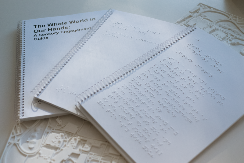 Documentation of the sensory engagement guide; three booklets are placed on table overlapping the top publication is opened to reveal a page with Braille, the bottom booklet show the title of the guide in lar print. They are stacked on top of two small tactile maps of UTM.