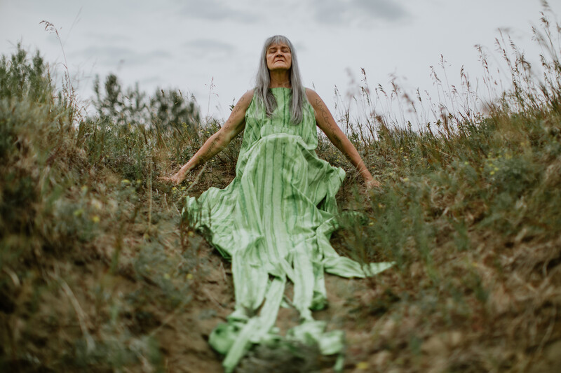 Seen from slighty below, Cheryl L’Hirondelle sits on a small bushy hill surrounded by prairie grasses. With closed eyes, her face points upward, and hands are outstretched to the ground on both sides. She wears a pale green dress with vertical stripes, and long strips of fabric cascade toward the camera. Her gray hair falls to the front, below her shoulders. Cree syllabic text is written on both of her arms.