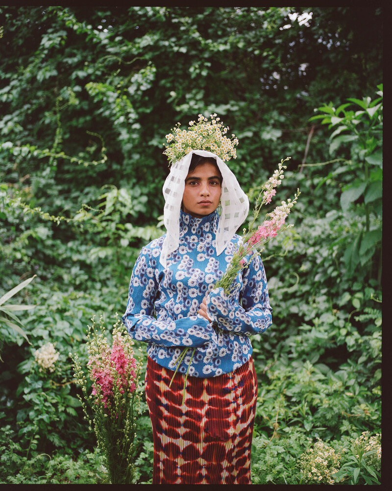 Róisín stands in a garden setting, holding a small bouquet in front of her chest, and with another cluster of small flowers perched atop her head, beneath a semi-translucent white scarf that falls to both sides of her face. She wears a blue dandelion-print turtleneck and a bright maroon and off-white tie dye-style skirt. The edges of the film frame are visible, and both sets of flowers she holds can be seen planted nearby.