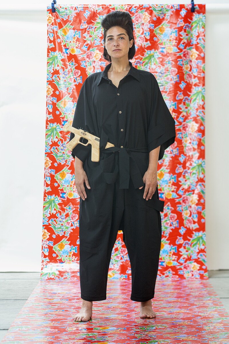 Standing in front of a colourful floral backdrop in red, blue, and green, Irmgard Emmelhainz wears a loose-fitting black jumpsuit, with an imitation wood machine gun tucked into her belt. She looks defiantly into the camera, with her hands hanging at her sides, and long hair swept back prominently over her head. The narrow backdrop is held up by two visible clamps, and it cascades down from a white wall in the background to the floor beneath her feet.