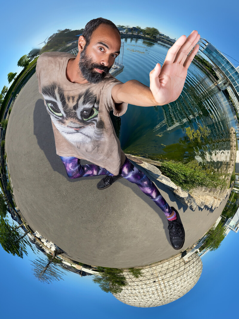 Seen from above, Luis Jacob stands with his left arm outstretched overhead, palm open, and his left leg pointed out at the same angle. He wears an oversize pale brown T-shirt with a large cat printed on it, and galaxy-print tights. He stands in front of a stylized panoramic image, which encircles him like a globe. The image is taken at Toronto’s Ontario Place, with a pond, trees, and geodesic dome visible at various points behind him.