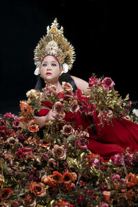 Partially leaned over to one side in front of a black backdrop, Melati Suryodarmo looks into the camera with a wide-eyed expression. She wears an elaborate gold Indonesian headpiece, white earrings resembling flower petals, and a red dress. In the foreground is an elaborate floral arrangement in orange, pink, and red, with some gold leaves. Suryodarmo also holds bouquets in both hands.