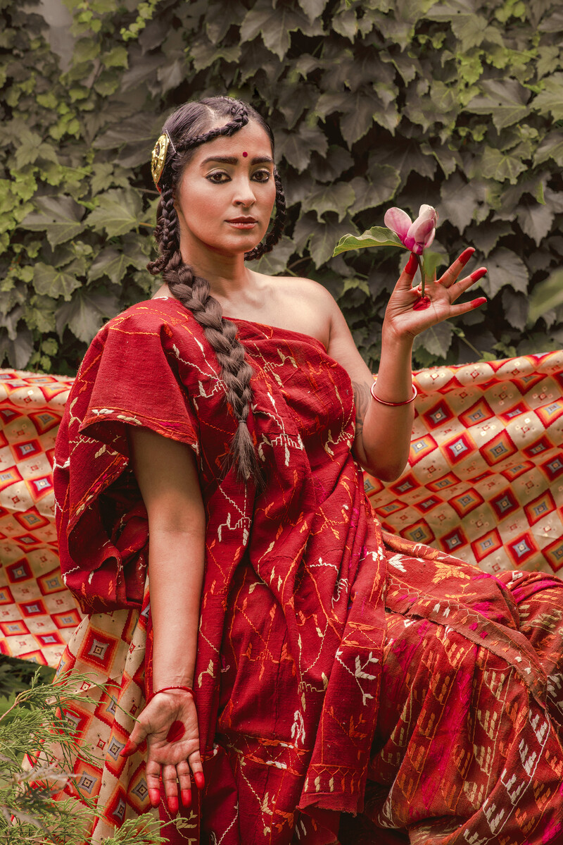 Raji Aujla sits facing the camera in front of a wall of greenery. She raises her left hand to shoulder height, delicately holding a lotus bloom. Her right arm points downward, with her palm facing the camera. She wears a red bindi on her forehead, larger circles painted onto her palms, and her fingertips are fully painted. Raji wears a red embroidered sari, and her braided hair falls in front of the right shoulder. Another patterned textile in red and beige is strung behind her across the bottom half of the image.