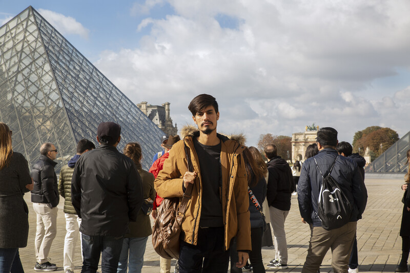 Ramin Mazhar is seen standing on a partly sunny day in front of the glass pyramid at the Louvre in Paris, France. He slings a brown backpack over his right shoulder, and looks into the camera without smiling. He has a mustache and goatee, wears a brown fur-lined jacket, and his hair is swept off to the left. Behind Mazhar, groups of tourists face away from the camera, standing loosely grouped as they listen to a tour guide.