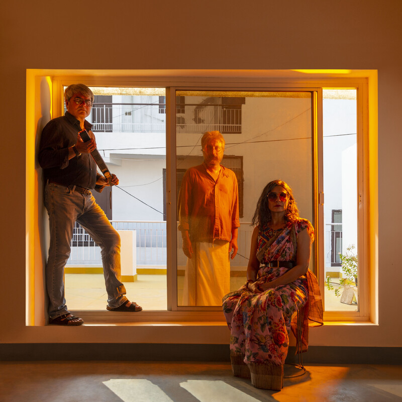 The three members of Raqs Media Collective (Monica Narula, Shuddhabrata Sengupta, and Jeebesh Bagchi) are situated inside and outside a glass sliding door, a looking at the camera. One stands in the threshold at left, holding a long, rectangular strip light in both hands. A second stands in the middle with hands limply at his sides, outside the doorway but visible through glass, wearing a translucent, dome-like mask. A third sits inside the room at centre-right, wearing a sari and large sunglasses. The room is cast with an orange glow, and the doorway backs onto a neighbouring apartment building.