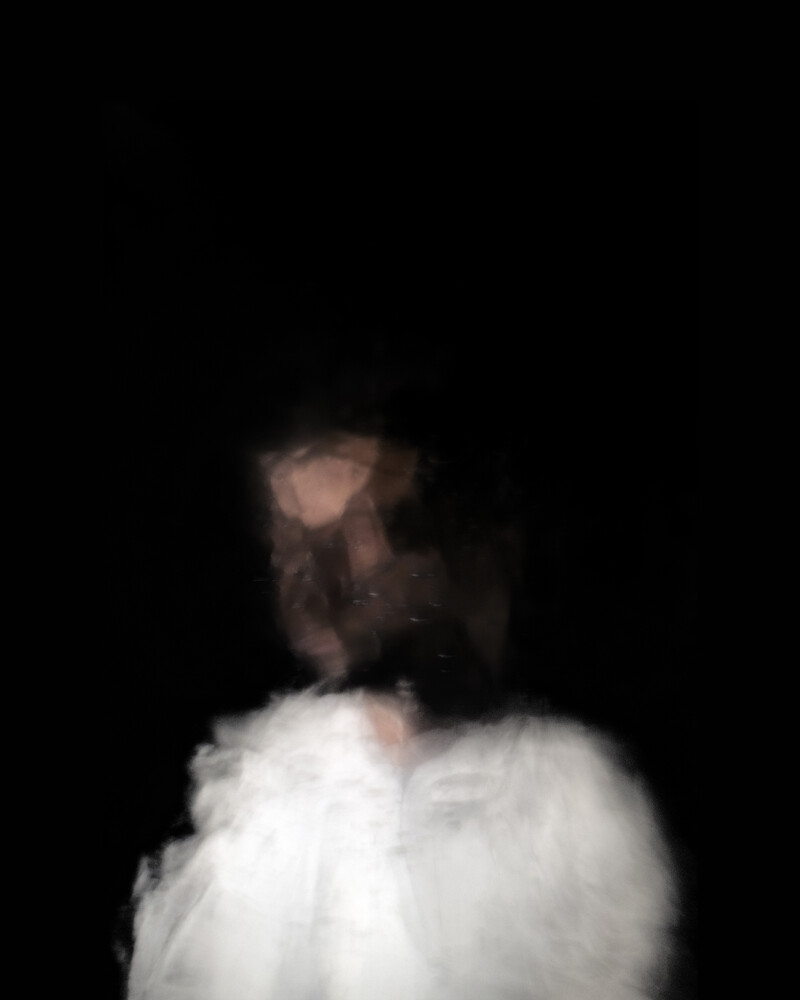 A person, presumably Roy Dib, appears indistinct in the lower half of the vertical-format image, in front of a black background. The image effect is blurry and kaleidoscopic, inhibiting the ability to see the subject. The outline of a person from mid-chest upward is visible, wearing a white shirt.