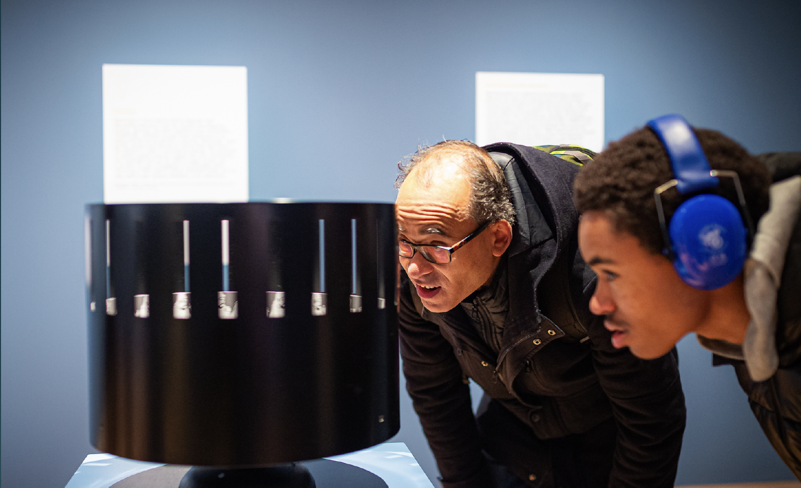 Two people bend over to look closely at the spinning zoetrope, a black steel circular device containing images inside that become animated when spinning. The individual on the far left wears blue earphones or ”ear defenders” for noise cancellation.