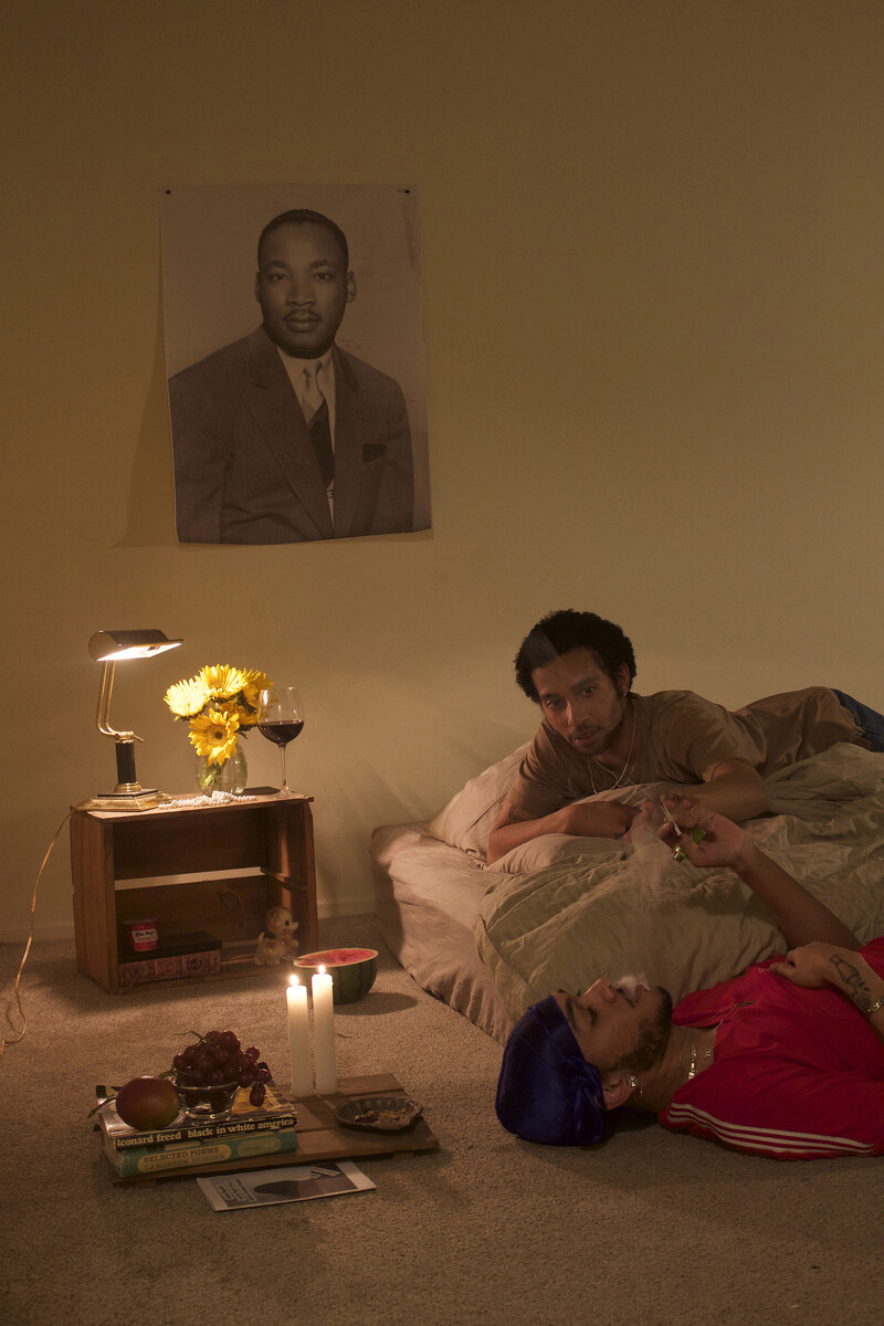 Two young Black men laying down, one on the floor looking up, the other on a mattress. They are sharing a joint, surrounded by various objects: a wine glass, watermelon, vase with yellow flowers, bowl of grapes, candles, and curios. Looking over the scene is a poster of Martin Luther King.