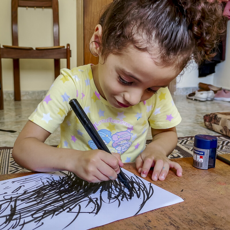 A young girl with curly brown hair tied up in a ponytail sits at a table holding a black marker, scribbling black lines across a white piece of paper.