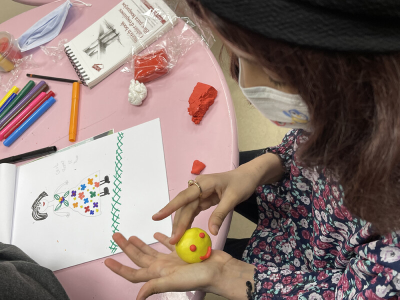 Photographed from above, a young girl wearing a mask and hat moulds yellow and orange clay into the form of a smiley face. On the table in front of her are various art supplies and a drawing of a girl wearing a flower-patterned dress in a field of grass.