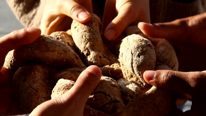 Three sets of hands holding a knotted loaf of bread in inconsistent lighting.