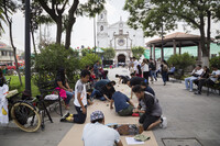 Youth gathered in a public square in Ecatepec drawing on a long sheet of kraft paper on a concrete surface, surrounded by trees and other foliage. People seated on benches and passersby curiously look on. A large white building with a bell tower and clock as well as a clay tile roof gazebo can be seen in the background.