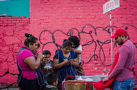 Situated against a pink painted brick wall with black spray-painted graffiti is a group of people consisting of men, women, and children gathered around a cart. The cart with a signpost that reads “El Carrito” in red and white lettering carries baskets and a tall metal pot. While some of the people write on small clipboards attached to the cart, others look on.