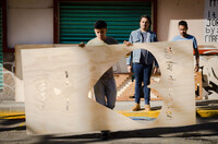 A man is shown in mid-stride, carrying a large wooden stencil with cut-out heart shape and cut-out text that reads; “dolor de corazon.” Two other men who look on from behind. All three men are situated along a street in the Mexican city of Ecatepec. The urbanscape depicted includes a dark green coloured garage door, details of a graffiti tag, and a sidewalk with yellow painted curb.