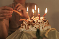 A baked alaska with lit birthday candles is displayed, elevated on a table covered with light green satin cloth. A figure in the background is shown inserting limp carrot sticks into the featured dessert.