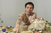 Centred in the frame is quori theodor who wears a white tank top, revealing tattooed arms, neck and chest. They are pictured against a grey background, at a table with many objects including: a baked alaska; a brown egg in a blue egg cup; a dish with fiddleheads; a stuffed sock; an iridescent conch filled with carrot sticks; and a glass cup filled with beige fabric and a green ribbon bow.