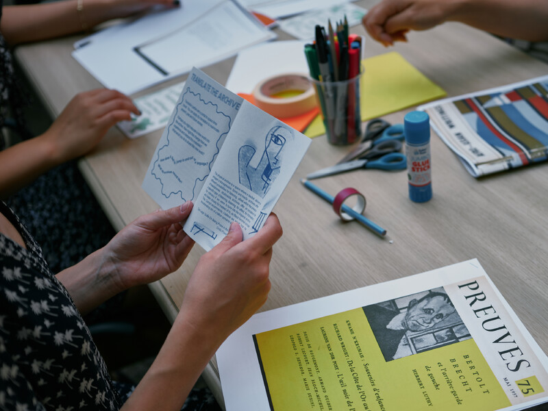 Close-up of workshop participants’ hands at a table with different printed materials, sheets of coloured paper, and an assortment of office supplies including pencils and markers, tape, a glue stick, and scissors. Prominent in the image is a set of hands holds a small booklet that opens to two pages featuring block text with a header that reads: “translate the archive.” There is also a colour-photocopy of the cover of Preuves Magazine, issue 75, printed in Mai 1957 featuring an artistic rendering of German playwright and poet, Bertolt Brecht.