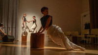 Centred in the foreground is a dancer wearing white pants and a dark-coloured top, leaning deeply and diagonally on a wooden block. In the background are two other dancers who also wear white pants and dark-coloured tops. Both dancers hold each other’s hands and both have one leg slightly bent.