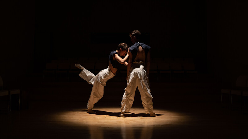 Two dancers are spotlighted on the dance floor as they partner; one dancer plants their feet in a stable stance while the other leans forward. They touch the shoulder of their partner and extend their left leg behind them.