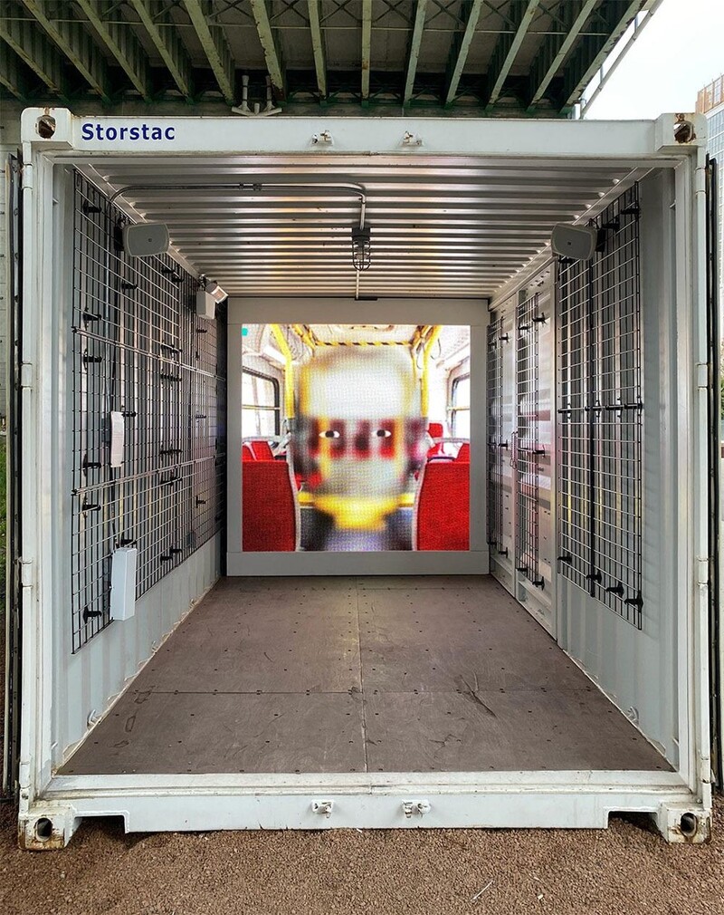 An image looking into a shipping container; on the end wall is a square projection of a face inside a public transit vehicle. The face has only cartoon-like eyes visible and is semi-transparent over the background, the result of being rendered in face-tracking software. 