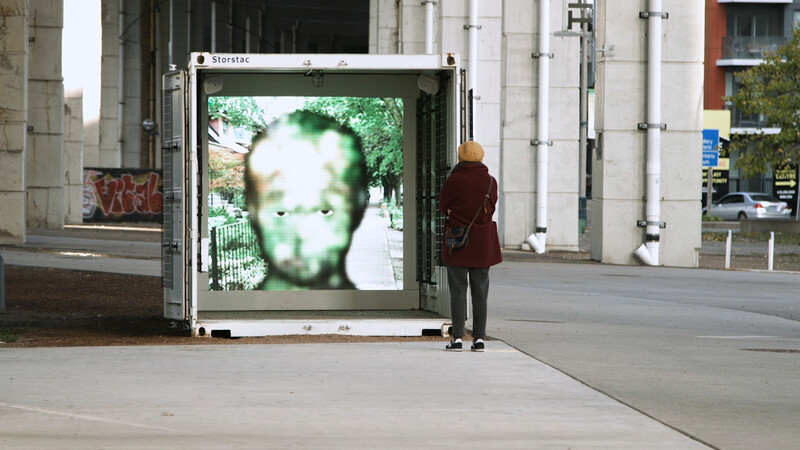 An image looking a shipping container with open doors, installed at an urban underpass in Toronto. On the end wall of the container is a square projection of a face in city space outdoors. The face has only cartoon-like eyes visible and is semi-transparent over the background, the result of being rendered in face-tracking software. 