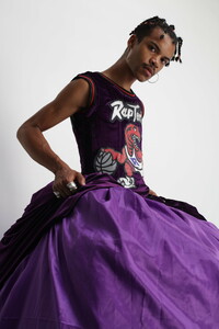 A single figure is seen looking directly in to the camera while posed for a tightly framed portrait. They are wearing short braids, large hooped earrings, large rings on their fingers and Mohamoud’s sculpture, a purple Toronto Raptors basketball jersey sewn into a large gown.