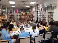 A large group of participants situated around three foldable tables pushed together partake in a zine workshop inside of The Beguiling bookshop. The group is working with a heap of craft and collage materials on the table, surrounded by books and bookshelves.