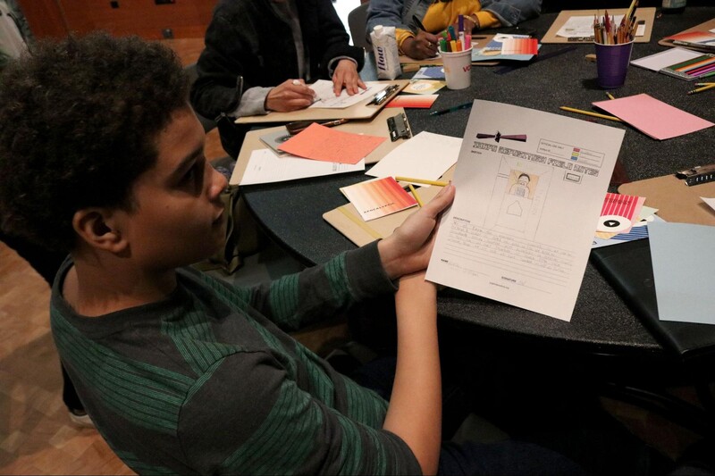 A workshop participant sits at a round table with a black tablecloth. The table has other participants working at it, and is covered in craft supplies, paper and coloured pencils for creating the manuscripts. The participant in focus holds their manuscript draft toward the camera.