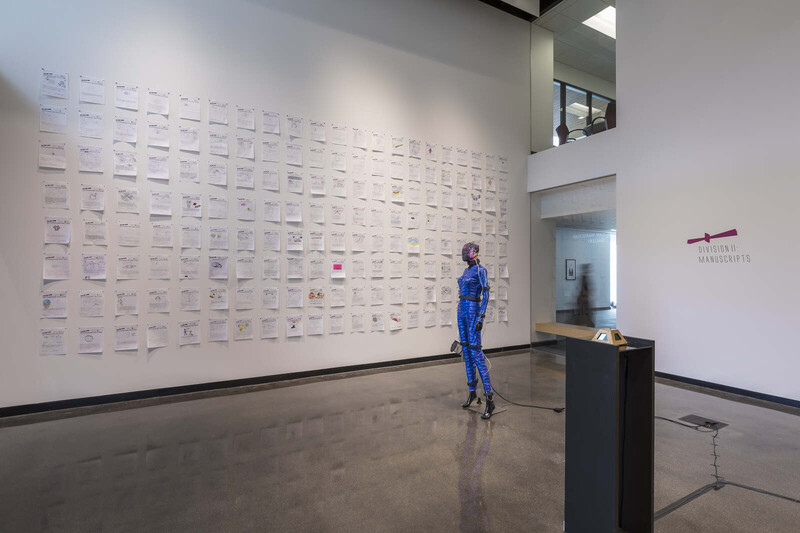 A gallery view of Iyapo Repository Manuscript Division installed at Law Warschaw Gallery. Paper manuscripts hung in a grid fill the entirety of the gallery’s back wall. A blue suit costume adorns a mannequin positioned in the middle of the gallery. 