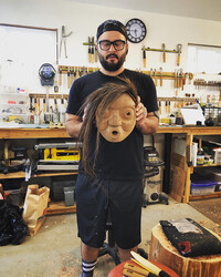 Standing in a woodworking shop, wearing a black t-shirt and shorts, Bracken Hanuse Corlett holds up a wooden mask center frame. A hair component is installed on the mask and falls over the left side of its face. The woodshop is decorated with organized tools in every direction of the background. 