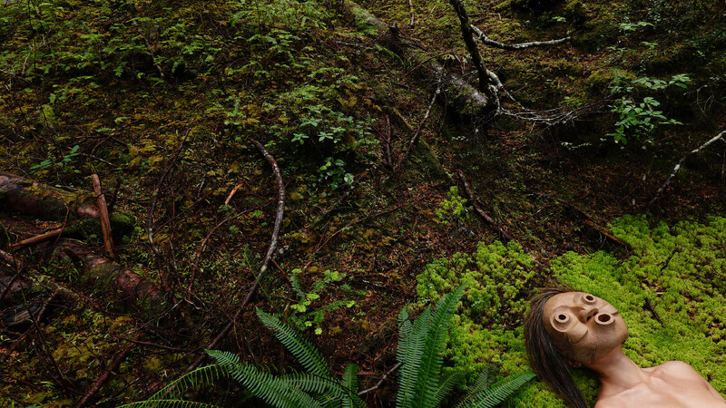 A green hilly landscape, with fallen logs, branches, small plants and ferns fill the entire image. In the very bottom right corner Lee Su-Feh is found lying in a bed of bright green moss. The mask covers her face as she looks directly up at the trees and sky.
