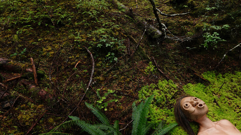A green hilly landscape, with fallen logs, branches, small plants and ferns fill the entire image. In the very bottom right corner Lee Su-Feh is found lying in a bed of bright green moss. The mask covers her face as she looks directly up at the trees and sky.
