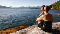 Lee Su-Feh is pictured sitting in a strapped dress holding the mask up to her face. She is sitting on a moss-covered rock overlooking the Pacific Ocean. Rock faces and mountains lined with coniferous trees enclose the body of water in the distance.