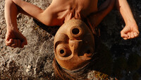 A tightly framed portrait of Lee Su-Feh laying against a rock on her back with clenched fists held up to her ears. The mask covers her face and she looks into the camera above.