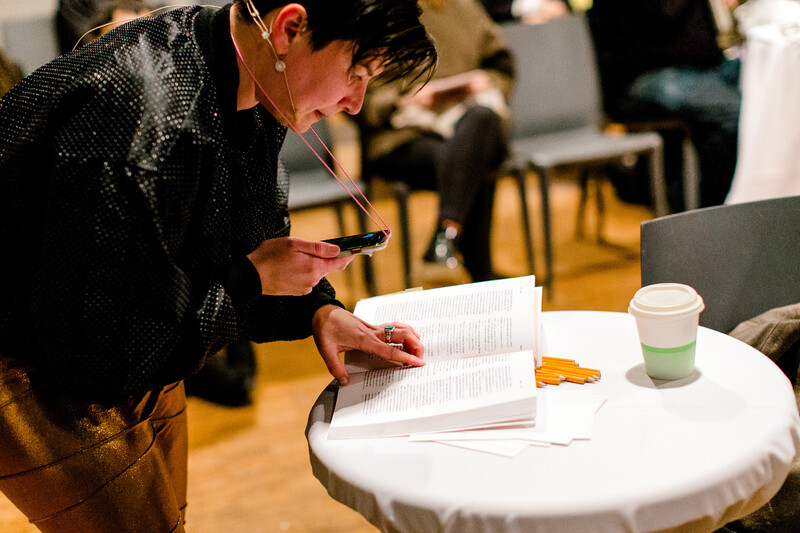 Vanessa is standing a in at the Harborfront Centre, with viewers sitting in chairs around her. She is leaning down on a table with a coffee and pencils on it, reading from the dictionary she performs with.&nbsp;She wears an iPhone on a necklace attachment around her neck, which is being used in the performance to record her reading from the dictionary. She wears a headset microphone and short dark hair.&nbsp;Shes also wearing a fabulous outfit, a pair of bronze sparkly pants, and a black silky bomber jacket with a spotted pattern all over it. 