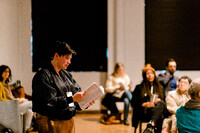 Vanessa is standing in a room at the Harborfront Centre, with viewers sitting in chairs around her as she reads from the dictionary she performs with in her hands. She wears an iPhone on a necklace attachment around her neck, which is being used in the performance to record her reading from the dictionary. She wears a headset microphone and short dark hair. Shes also wearing a fabulous outfit, a pair of bronze sparkly pants, and a black silky bomber jacket with a spotted pattern all over it.