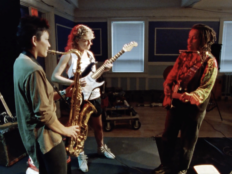 Faith Nolan (at right) faces two bandmates during a rehearsal in a community hall. Faith is illuminated in red, holding an electric guitar and with a harmonica around her neck; her bandmates play electric guitar and saxophone. Music gear is seen on the floor behind them.