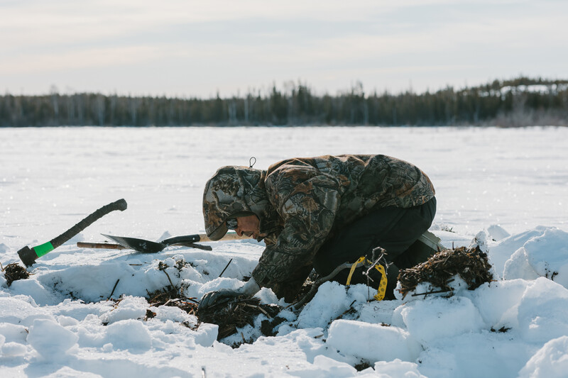 A person in a camoflauge patterned coat reaches into the ice. Snow blankets the land around them; an axe, shovel, and other tools scatter the snow.