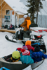 A person wearing a bright orange vest over their winter coat drives a ski-doo. They look over their shoulder at the camera. The ski-doo tows a carriage behind it; children bundled in winter hats sit inside.