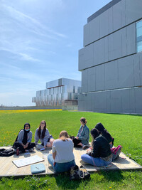 A group of people sit on a wooden platform in a green dandelioned field, with towering grey building behind them. Books are scattered around them and some people smile into the camera; they look like they are studying.