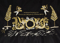 Gold threads embroider a mythical scene on black fabric: angels playing lyres fly in the air, and two humanoid figures with wings and mermaid tails hold a wreath in the centre. Trees, flowers, and swimming fish surround the scene. Text in the middle reads, 